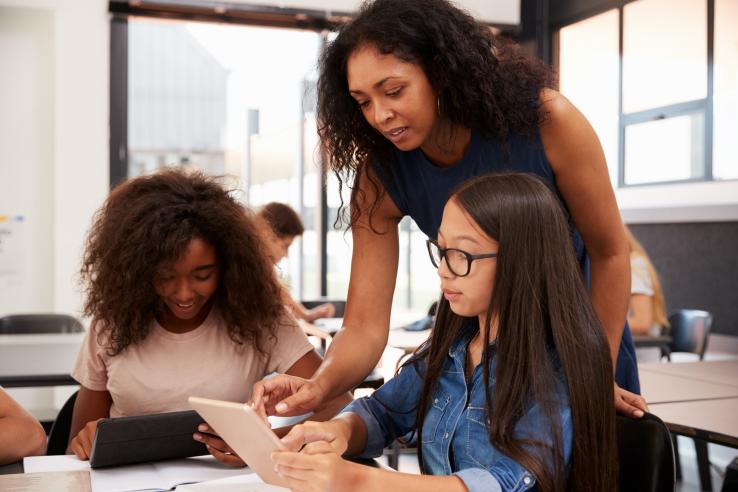 A teacher helps two high school students with technology in a classroom.