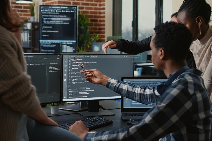 A man points to code on a desktop computer with two women