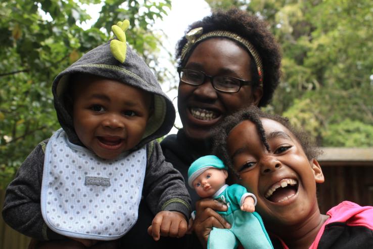 A Black mother laughs with her infant boy and young daughter holding a baby doll outside behind trees.