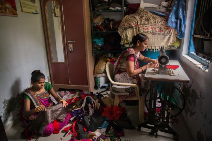 Two women are pictured surrounded by colorful fabric scraps. One women is sitting at the desk working with a grey sewing machine, while the other woman is sitting behind her cross legged on the floor folding a scrape of brown fabric.