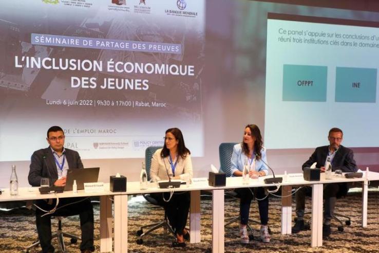 Four speakers are seated at a panel table at the front of a room. 