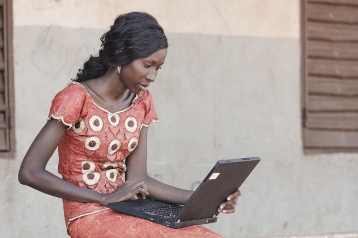 A woman wearing a red dress sits outdoors holding a laptop on her lap. 