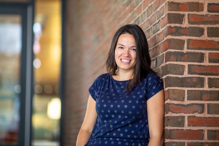 Elizabeth Linos is smiling for the camera in a short sleeve blue shirt standing against a brick wall