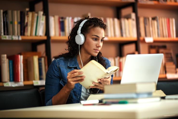 A woman wearing headphones and holding a book studies in a library and looks at a laptop.