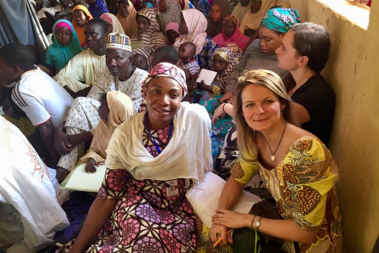 Two women sit on the floor and look at the camera, with a room full of people behind them.