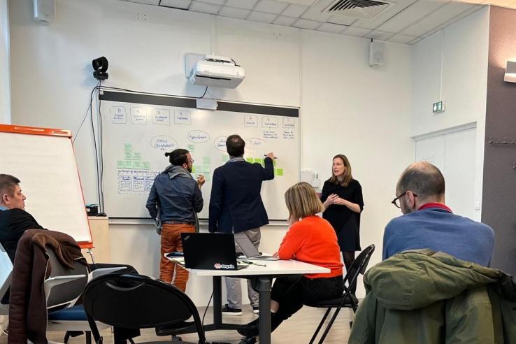 A group sits at tables while two people stand facing a white board.