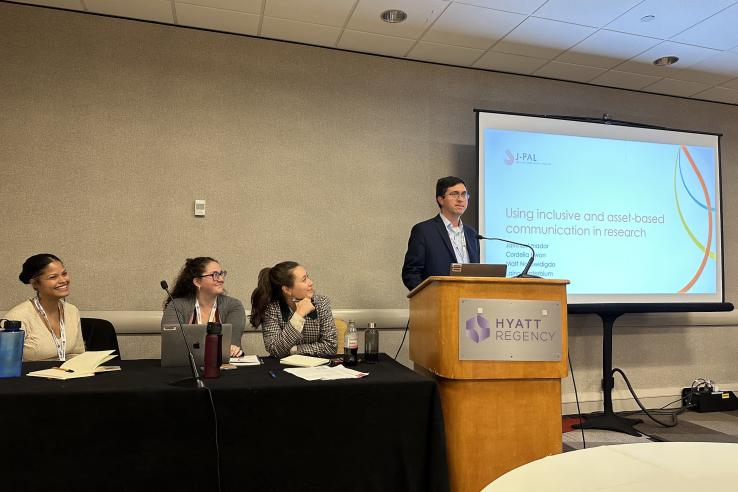 J-PAL staff (left-to-right) Jatnna Amador, Laina Sonterblum, and Cordelia Kwon sit at a table while J-PAL North America Co-Scientific Director Matt Notowidgdo stands at a podium next to a slide show that reads “Using inclusive and asset-based communication in research.”