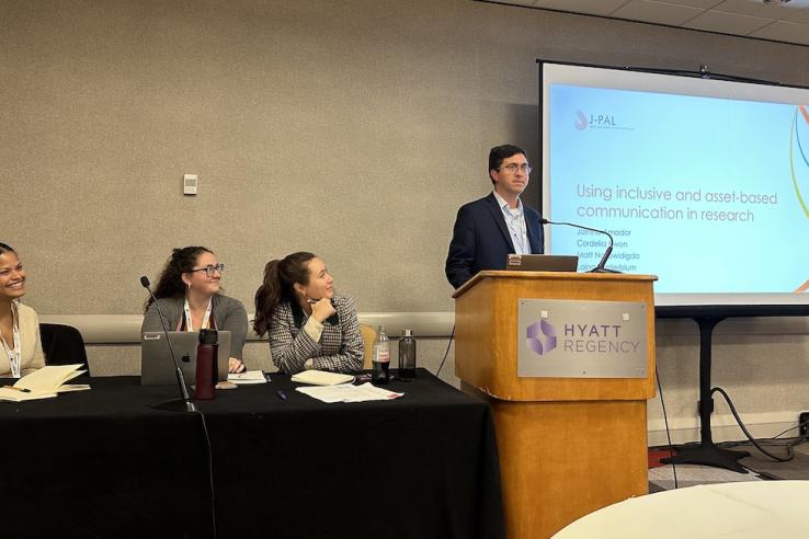 J-PAL staff (left-to-right) Jatnna Amador, Laina Sonterblum, and Cordelia Kwon sit at a table while J-PAL North America Co-Scientific Director Matt Notowidgdo stands at a podium next to a slide show that reads “Using inclusive and asset-based communication in research.”