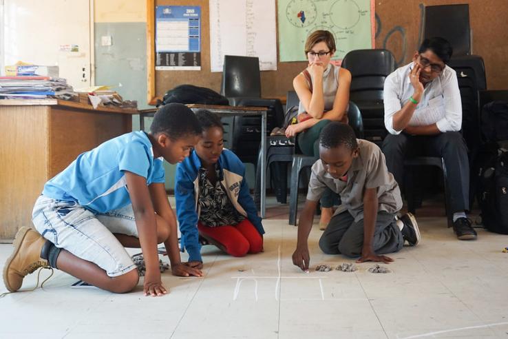 three children play on the ground while two seated adults watch