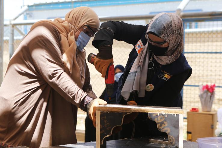 Two women wearing headscarves use a drill to upcycle a piece of wood furniture. 
