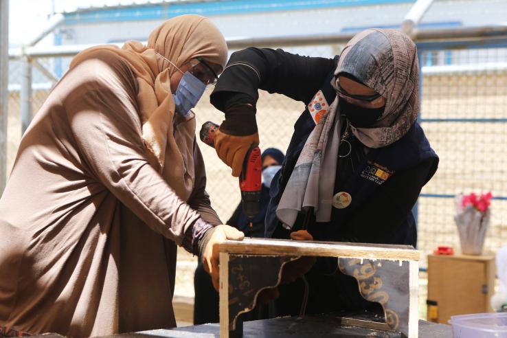 Two women wearing headscarves use a drill to upcycle a piece of wood furniture.