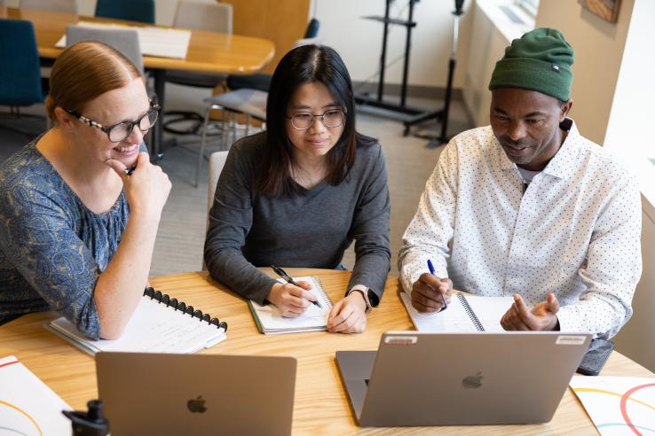 Three students sitting around a table with two laptops in a classroom.