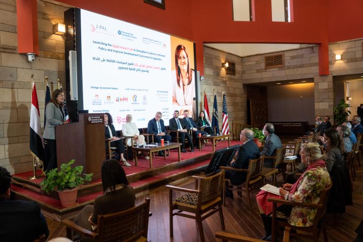 A group of people seated in an audience with their backs to the camera are looking at a stage on which a speaker is presenting a slide show about the launch of the Egypt Impact Lab. 