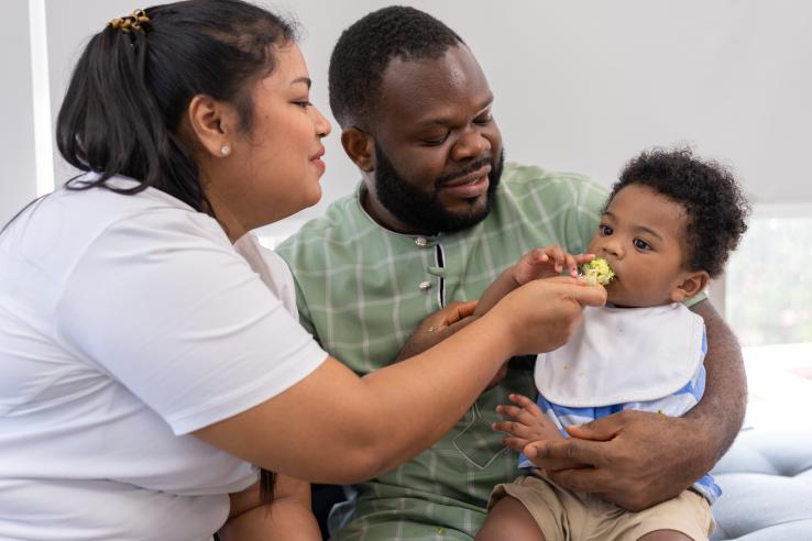 A woman is feeding her child while he is being carried by his father