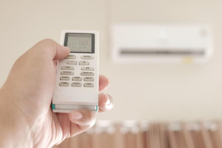 A person holds a programmable thermostat in front of an air conditioning unit.
