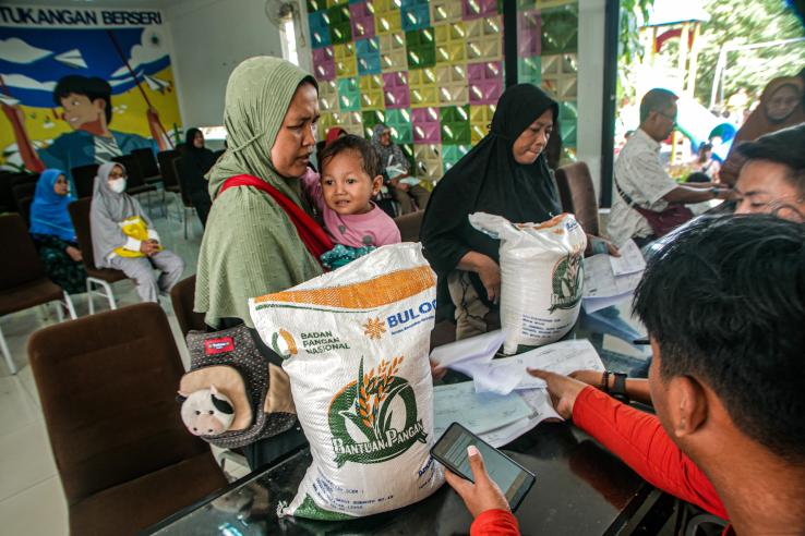 A woman wearing a green head scarf stands holding a child. In the foreground is a counter with a bag of subsidized rice.