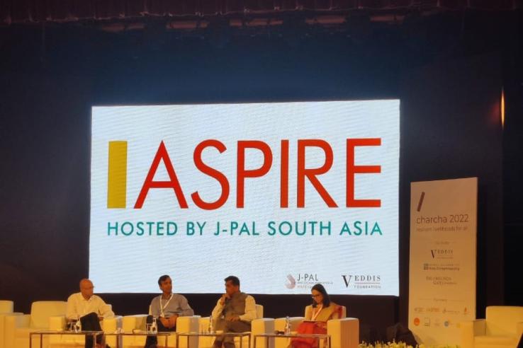 Four panelists seated on stage during Charcha 2022, with a large screen behind them displaying the ASPIRE logo and the text “Hosted by J-PAL South Asia.”