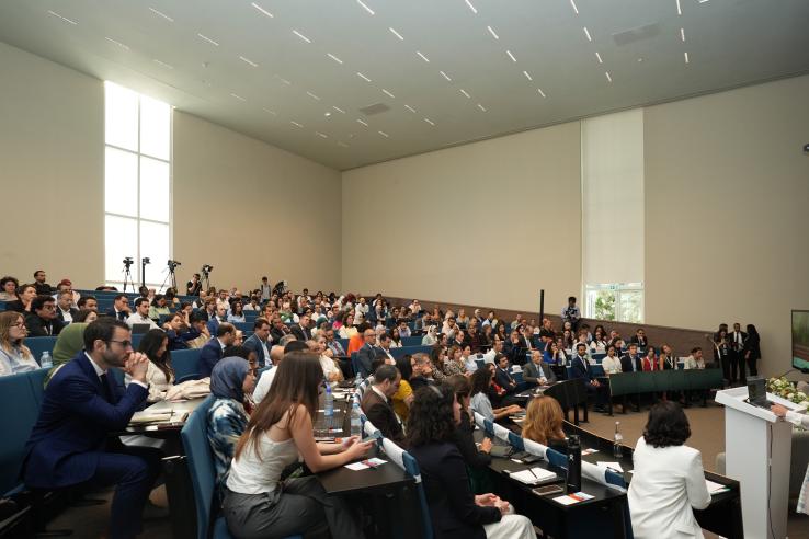 An auditorium filled with people facing a female presenter.