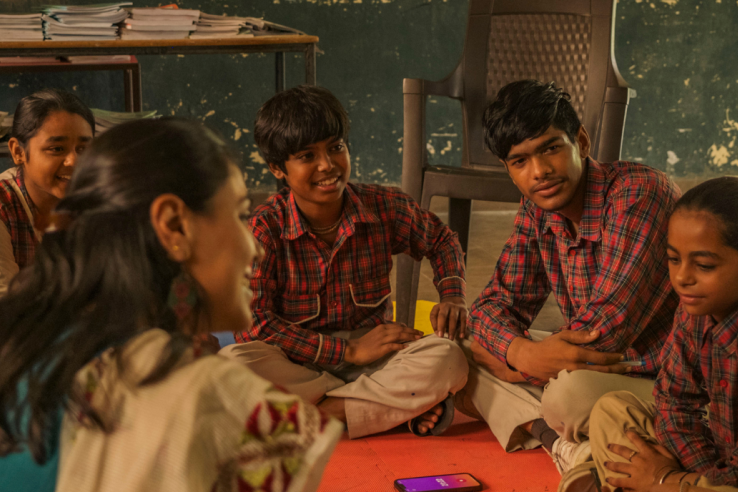 A group of schoolchildren in uniform sit in a circle on colorful mats, attentively listening to a woman facilitator in a classroom with a chalkboard and scattered learning materials.
