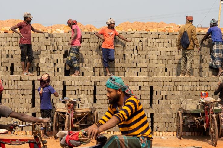 Bangladeshi traditional kiln workers stack bricks and converse with other workers in the background. In the foreground, other workers transport brick loads with bicycles. 