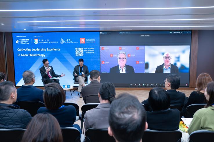 An audience watches several live speakers and two speakers on zoom