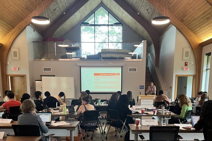 Students sit in a sunlit classroom in front of a lecturer and projected presentation.