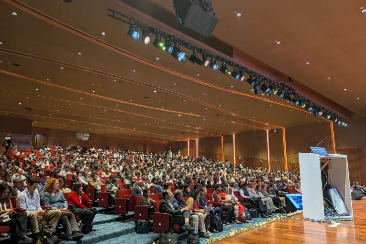 People sit in a large audience in a wood paneled room with a corner of the stage visible.