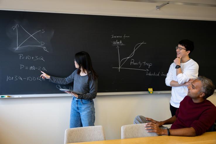 Student explaining economics equations on a chalkboard.