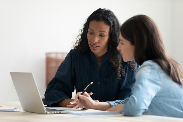 Two woman look at a laptop screen and take notes