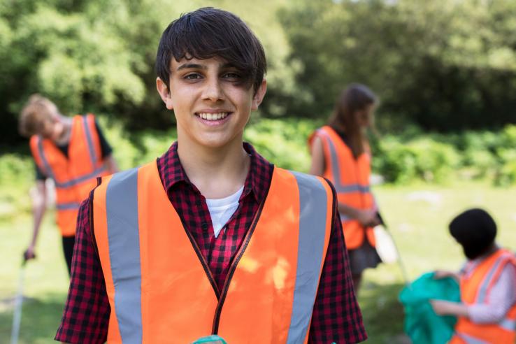A young person wearing a high-visibility vest smiling at the camera. Three individuals are behind them cleaning up a park.