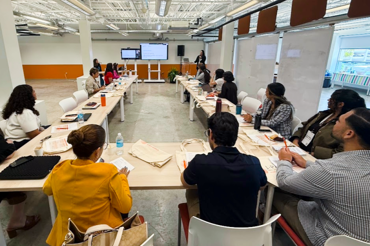A group of people sitting at a U-shaped table listening to speakers at the front of the room.