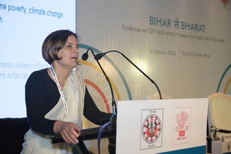 A woman in a black top and cream handloom saree speaks at a podium with two microphones, in front of a “Bihar se Bharat” event backdrop and a projected slide.