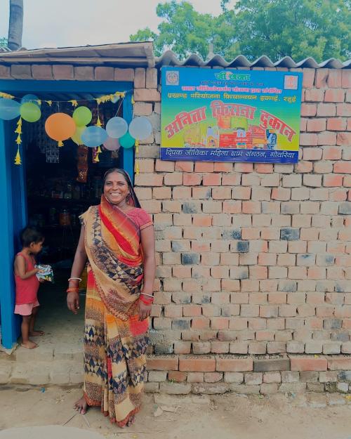A smiling woman in a gold and red sari stands outside her newly opened kirana (grocery) store, decorated with balloons, with a signboard reading "Anita Kirana Dukan" and JEEViKA branding above; a young child stands in the doorway holding a snack packet.