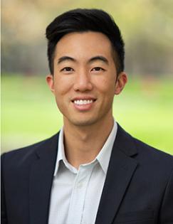 A head shot of Vincent Quan in a suit, standing outside smiling behind a grassy background.