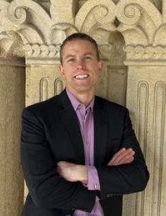 A man wearing a black blazer and red shirt stands smiling with his arms folded against a tan wall.