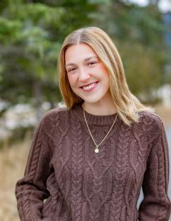 A girl stands smiling in a brown sweater
