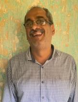 A middle-aged man with salt-and-pepper hair and a moustache is smiling warmly. He is wearing glasses and a grey checkered shirt, standing in front of a textured wall with peeling pastel green and beige paint.