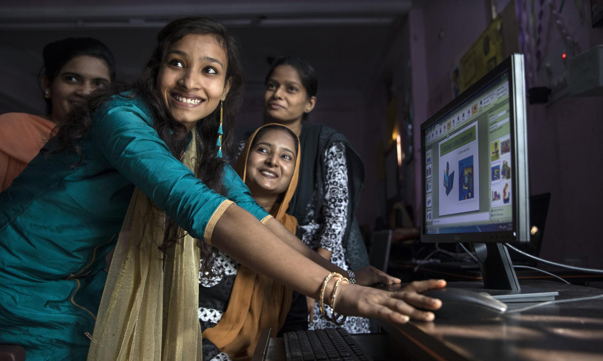 A group of four Indian woman stand in front of a computer and smile at something off-camera