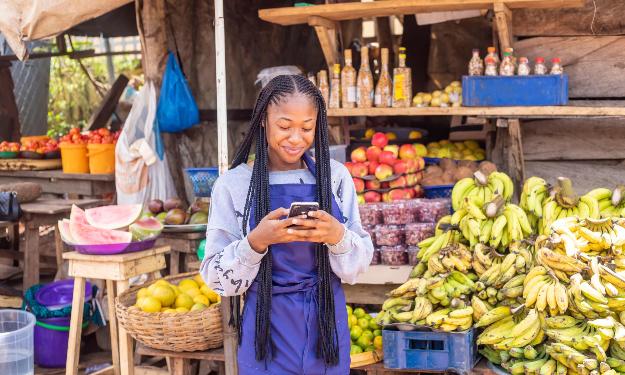 Young entrepreneur smiling while looking at her phone in front of her fruit stand