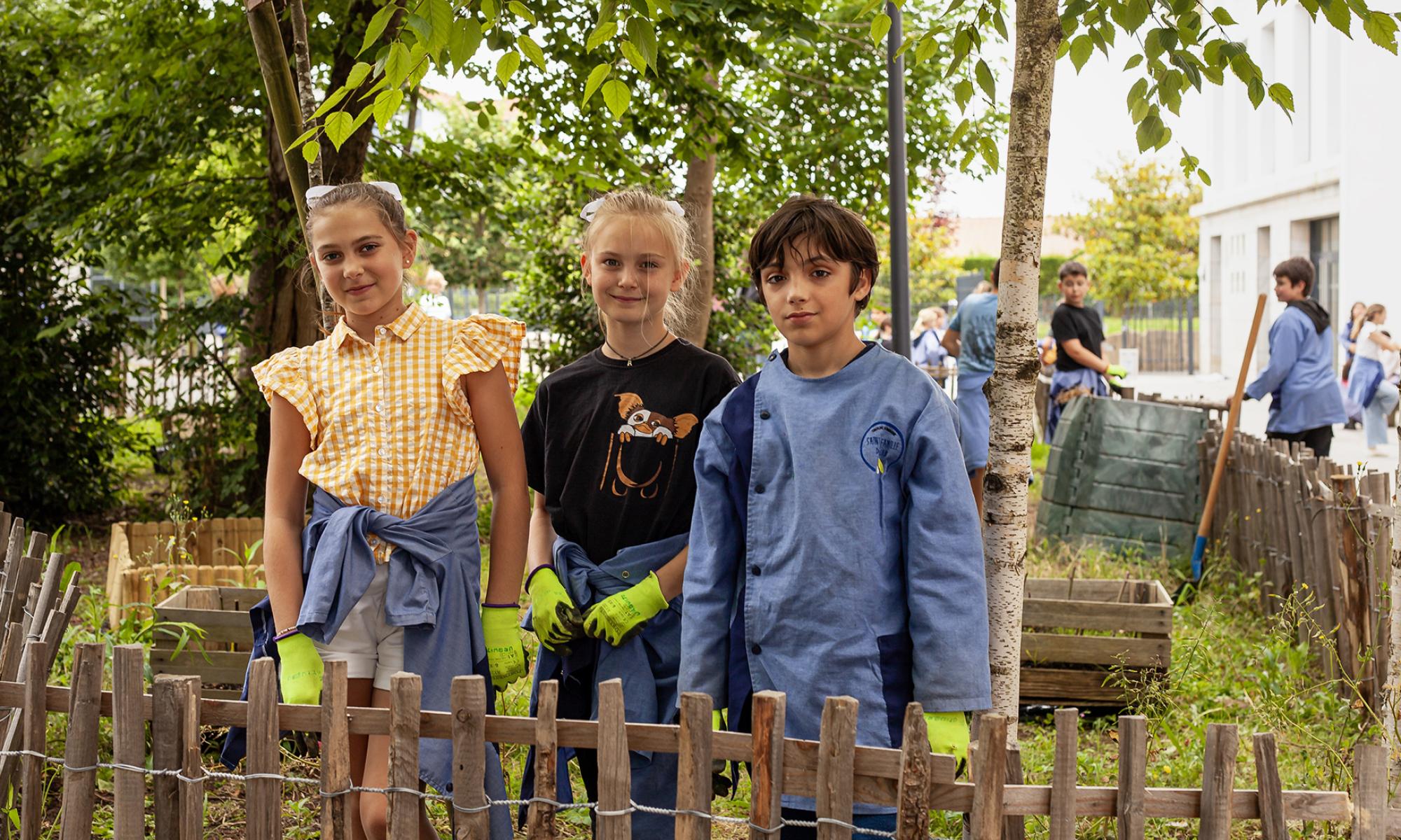 Two young girls and a boy stand with gloves on in a garden