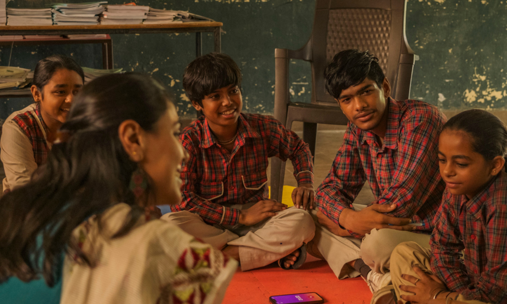 A group of schoolchildren in uniform sit in a circle on colorful mats, attentively listening to a woman facilitator in a classroom with a chalkboard and scattered learning materials.