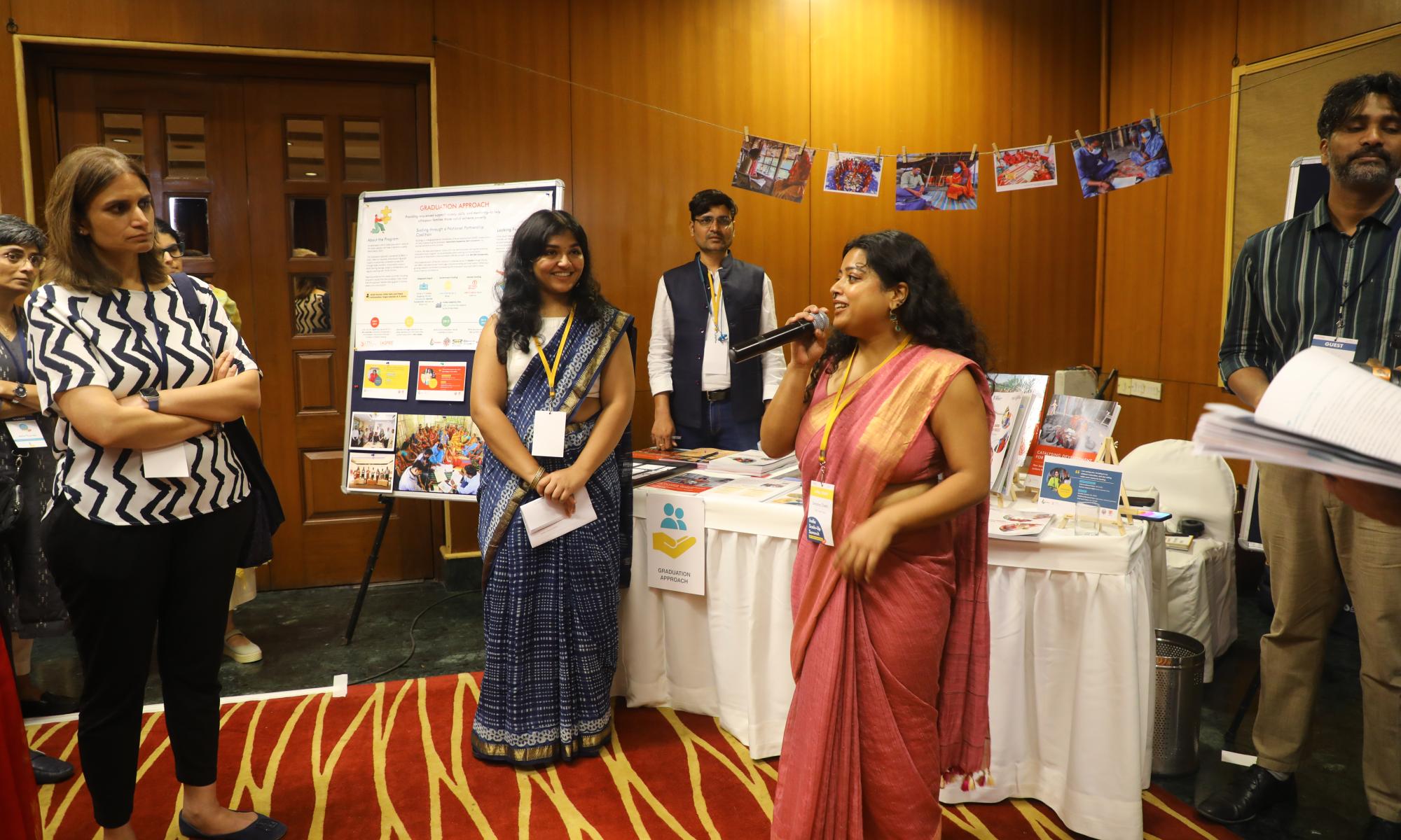 A woman in a pink sari speaks into a microphone while addressing a small group at an indoor event booth. Behind her, posters, photos, and materials are displayed on a table and board labeled “Graduation Approach.” Several attendees listen attentively, including a woman in a blue sari and another in a black-and-white patterned top. The setting appears to be a conference or exhibition with warm lighting and wooden paneling.