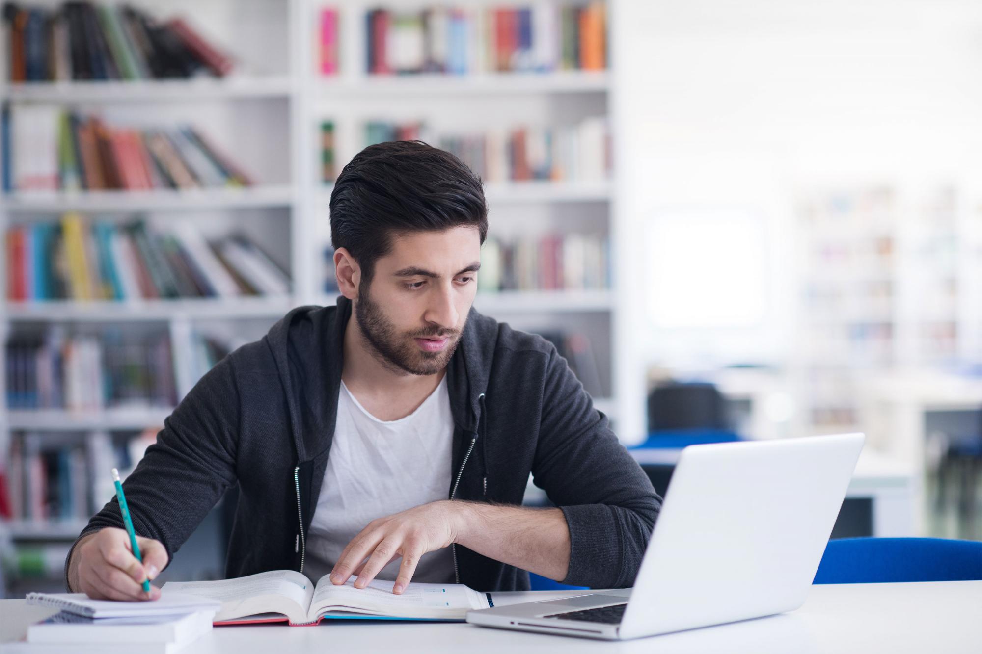 A man sits at a desk, looking at laptop screen while writing notes