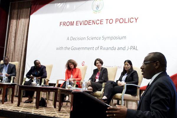 In the foreground, a man speaks at a podium. In the background, panelists sit on a stage.