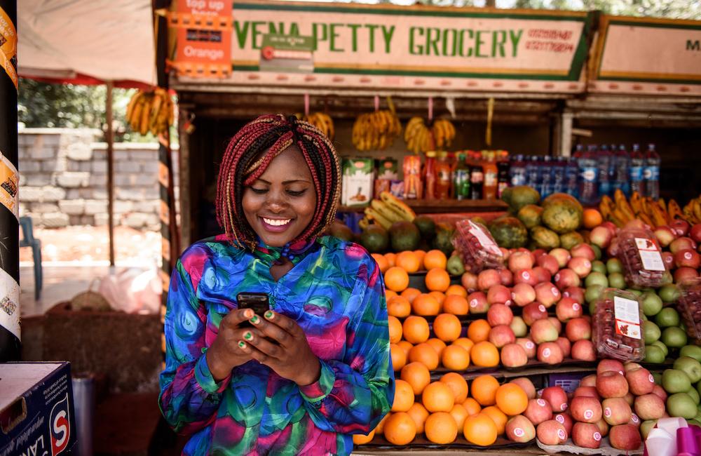 A woman looks at her phone while standing in front of a colourful fruit stand.