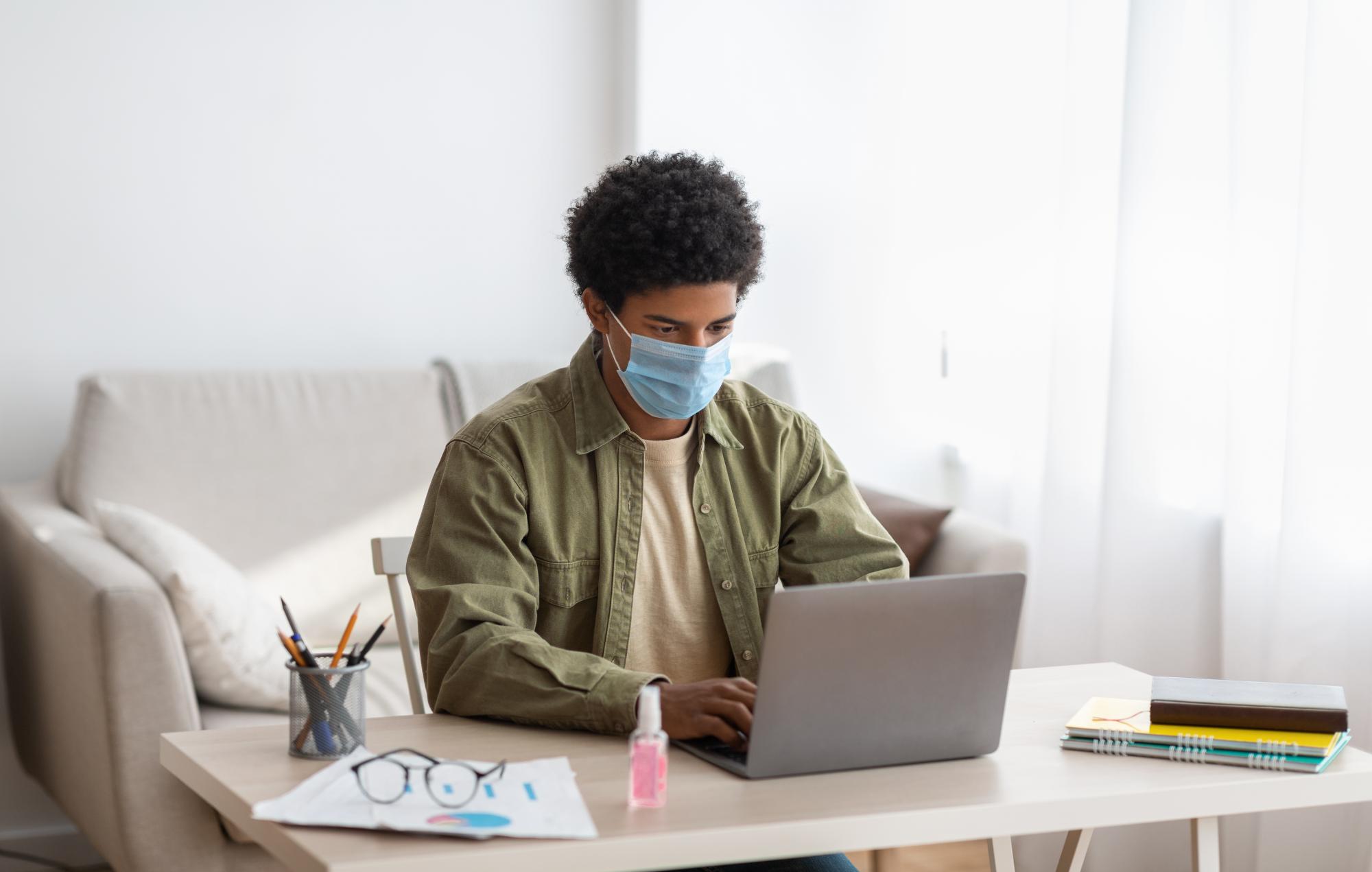 A man wearing a mask sits at a desk typing on a computer.