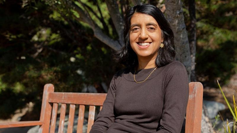 Seema Jayachandrani sits on a wooden bench outside and smiles at the camera.