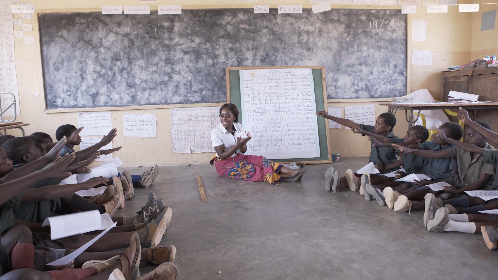 A teacher sits on the floor by a chalk board. Students on either side of her raise their hands.
