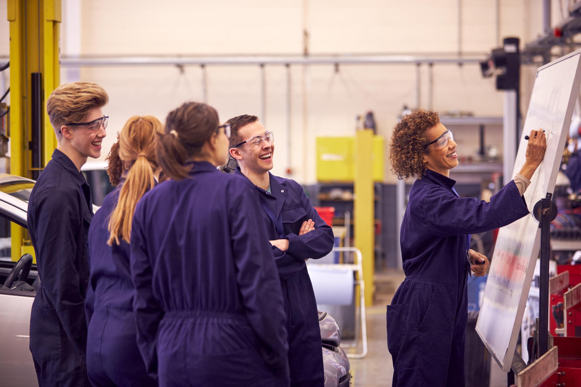 A group of workers in blue jumpsuits; one writing on whiteboard