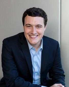 Headshot of Joshua Dean wearing a blue shirt and black coat